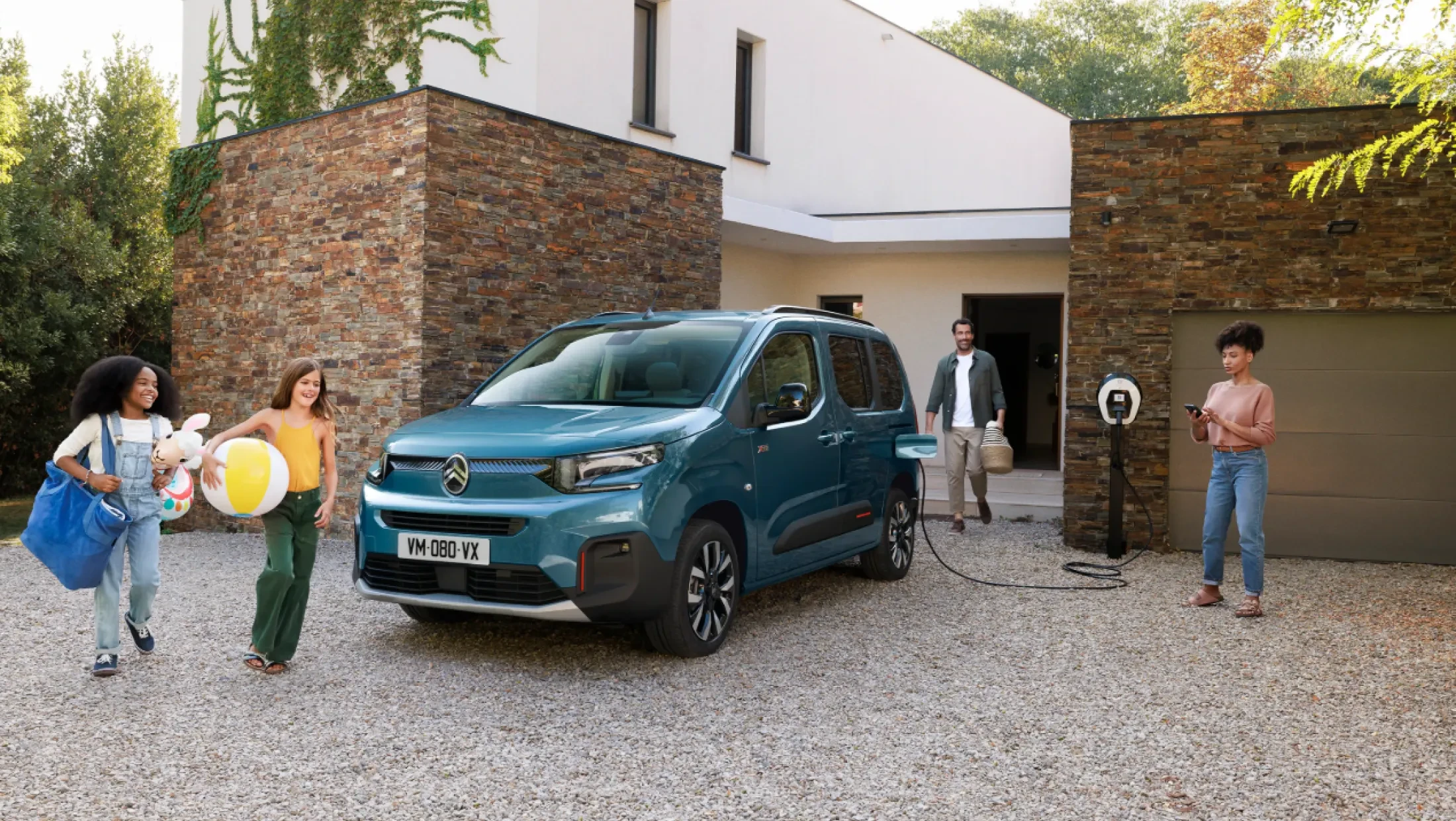 Voorzijde van de Citroën ë-Berlingo, aangesloten op een laadpaal, geparkeerd voor een huis met mensen die naar de auto lopen.